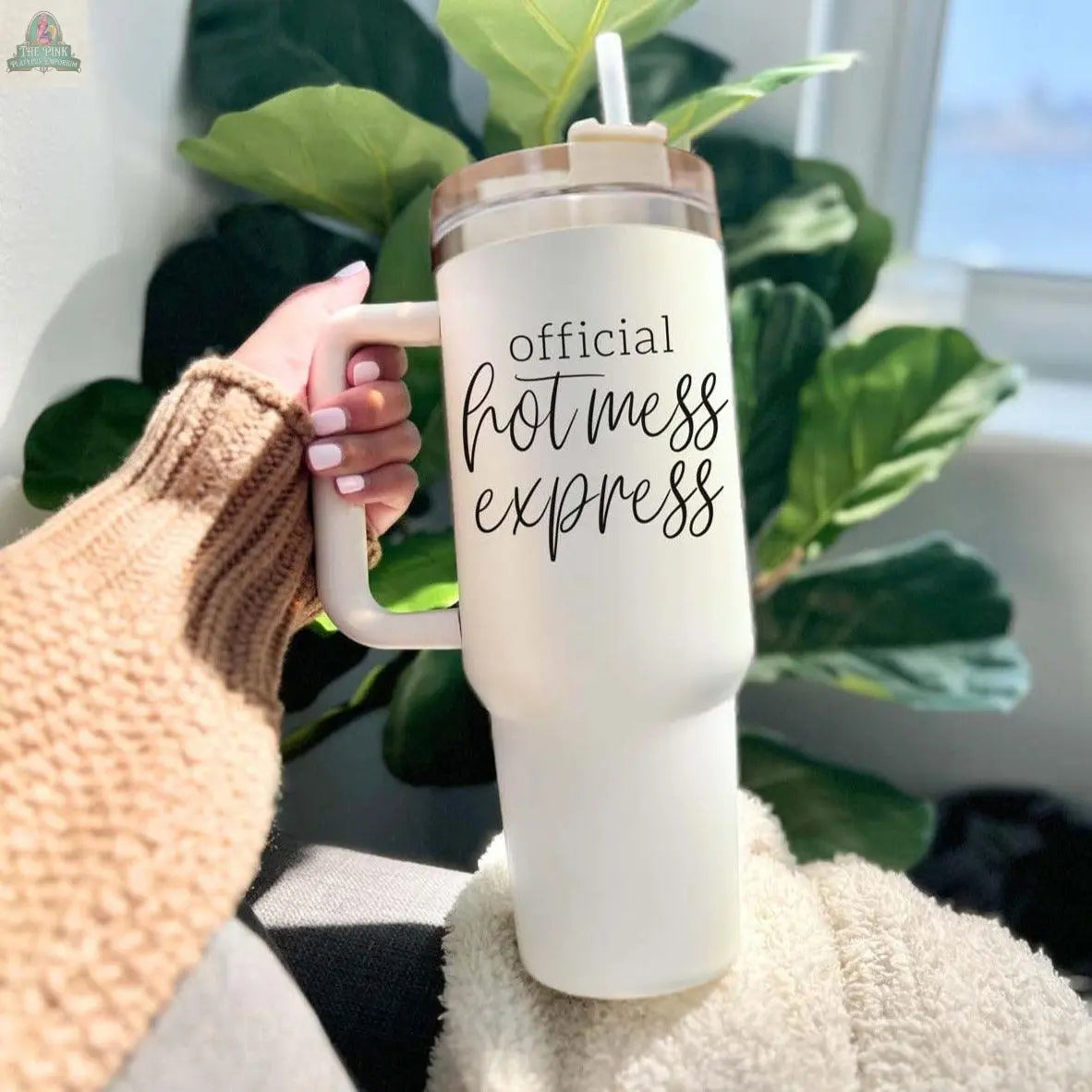 A hand holds the Official Hot Mess 40oz tumbler with a straw. The insulated cup features “official hot mess express” in bold black script, while green leaves and sunlight brighten the background.