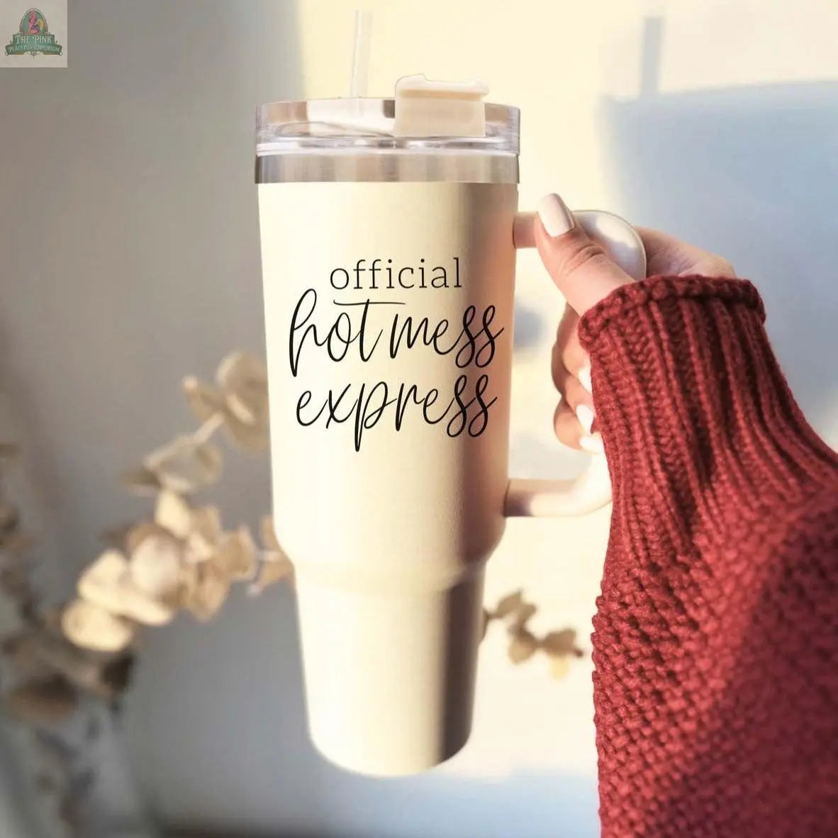 A hand in a maroon knit sweater holds the 40oz "Official Hot Mess" insulated tumbler with a straw, featuring black script. Light and dried flowers create a soft background.