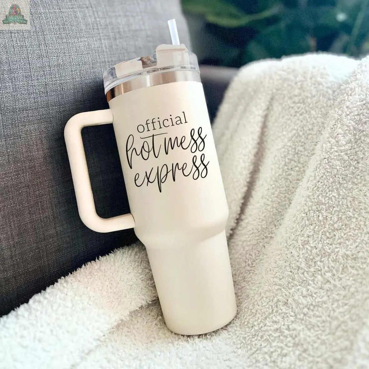 The Official Hot Mess 40oz tumbler in cream sits on a white blanket, featuring playful black script, a handle, and straw. This insulated travel mug is perfect for adventures and makes a fun gift.