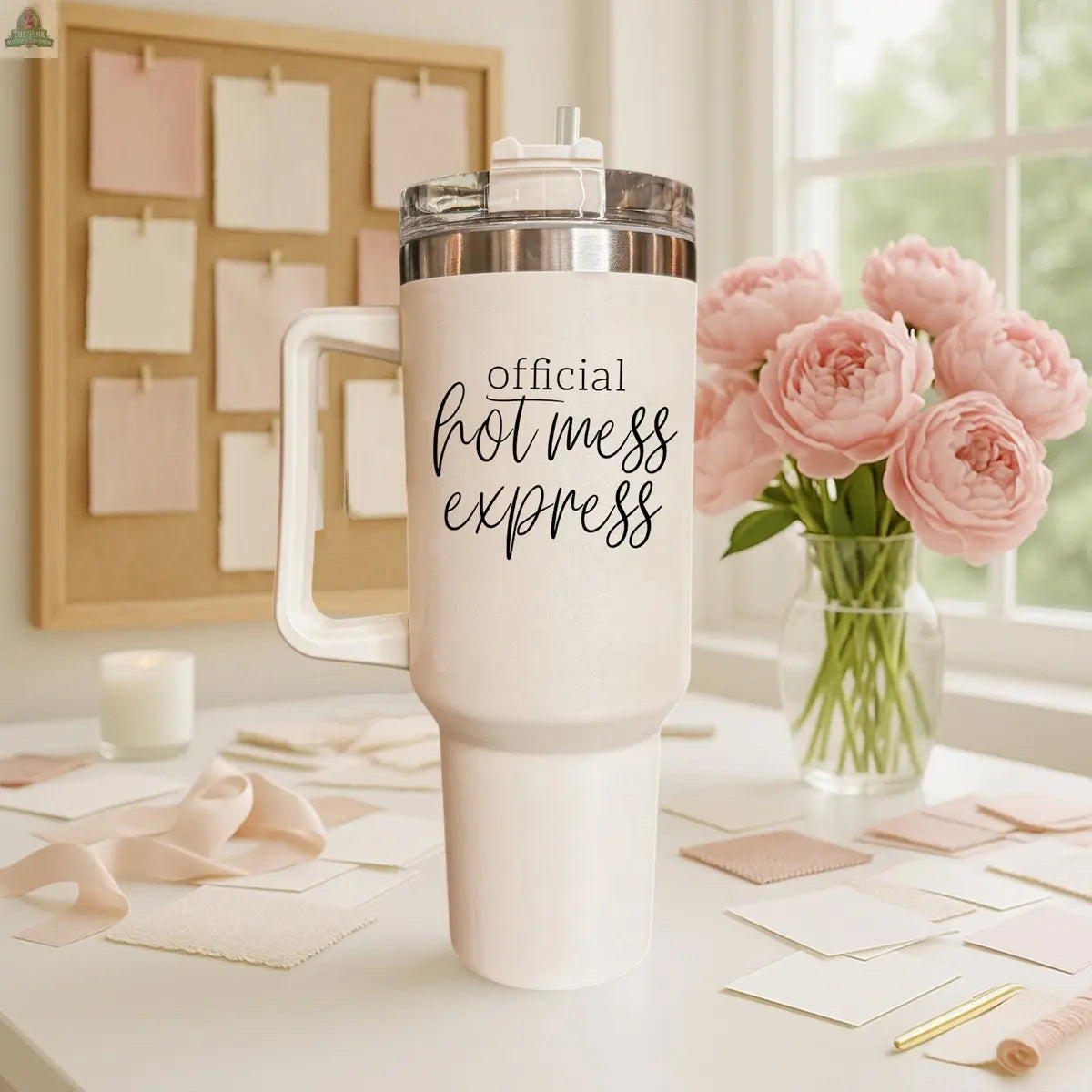 An Official Hot Mess 40oz insulated tumbler sits on a desk with pink flowers, fabric swatches, and a corkboard in a softly lit room.