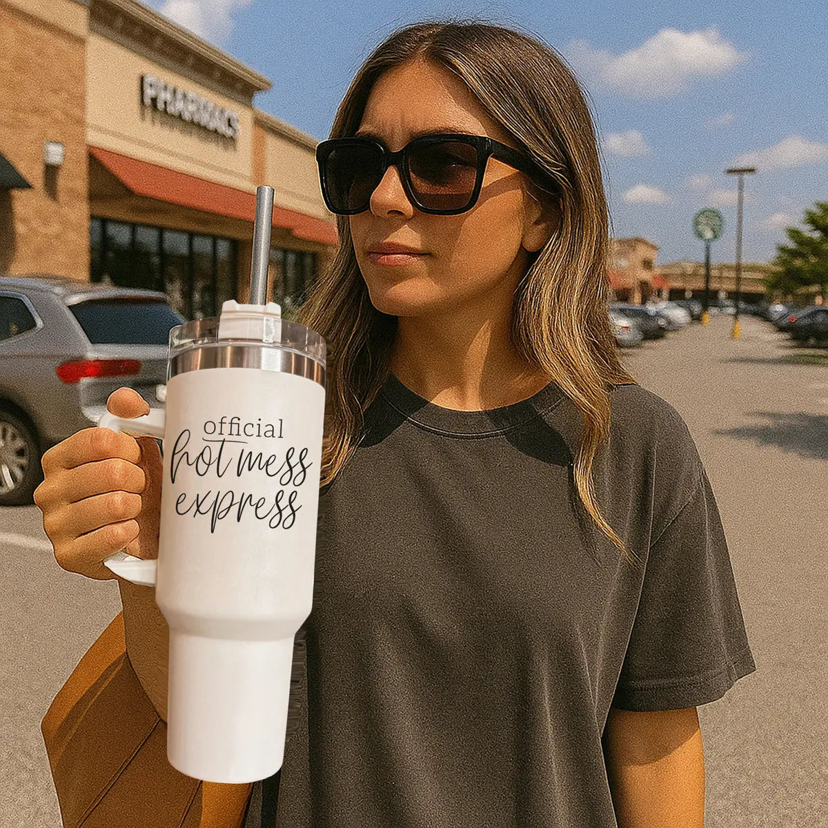 A woman in sunglasses and a dark t-shirt stands in a sunny parking lot, holding an Official Hot Mess 40oz tumbler. Cars and stores are visible in the background.