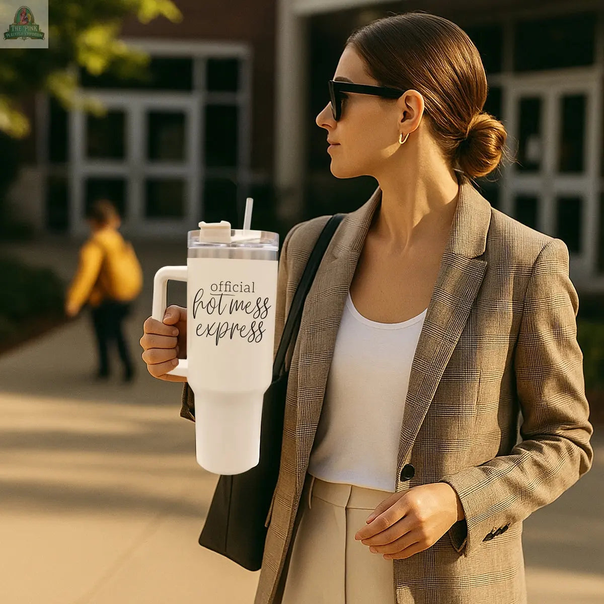 A woman in sunglasses and a plaid blazer carries the Official Hot Mess 40oz tumbler while walking outdoors; in the background, a child with a yellow backpack appears blurred.