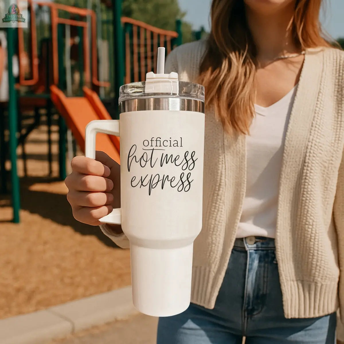 A woman at a playground holds the Official Hot Mess 40oz insulated tumbler with a straw. She wears a cream cardigan, white shirt, and blue jeans.