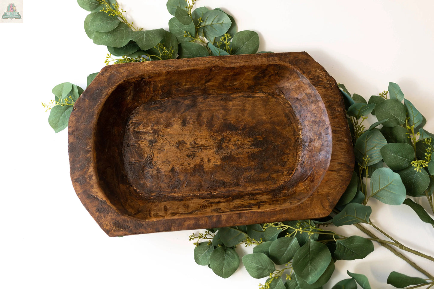 The Petite Wood Bowl (9-10"), handmade in Mexico from natural wood, sits empty on a white surface and is surrounded by green leafy branches for decoration.