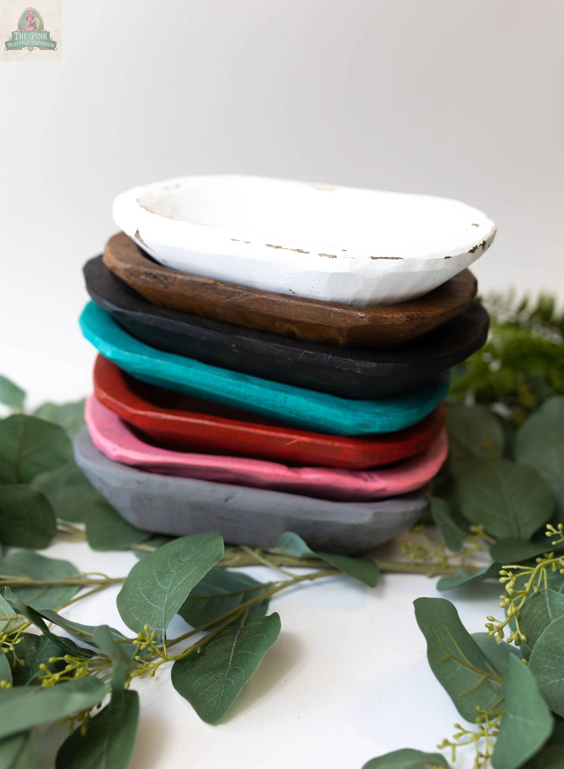 A Petite Wood Bowl (9-10"), handmade in Mexico from natural wood (10x6x3), sits on a white surface, surrounded by green leaves and small yellow buds.