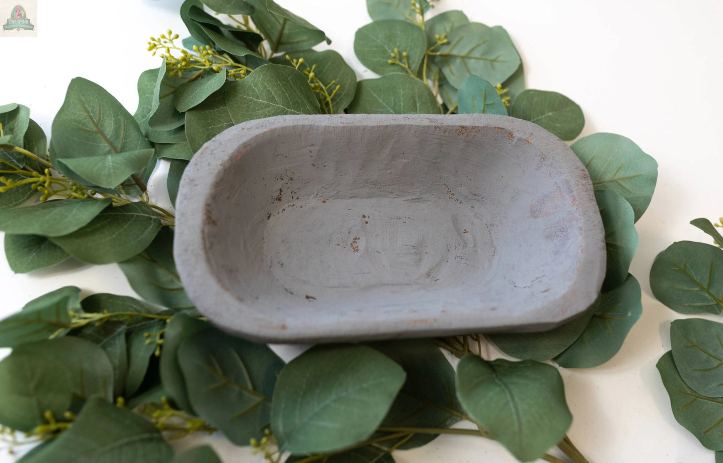 A handmade Petite Wood Bowl (9-10"), natural finish, 10x6x3 inches, from Mexico, sits empty on overlapping green eucalyptus leaves atop a white surface.