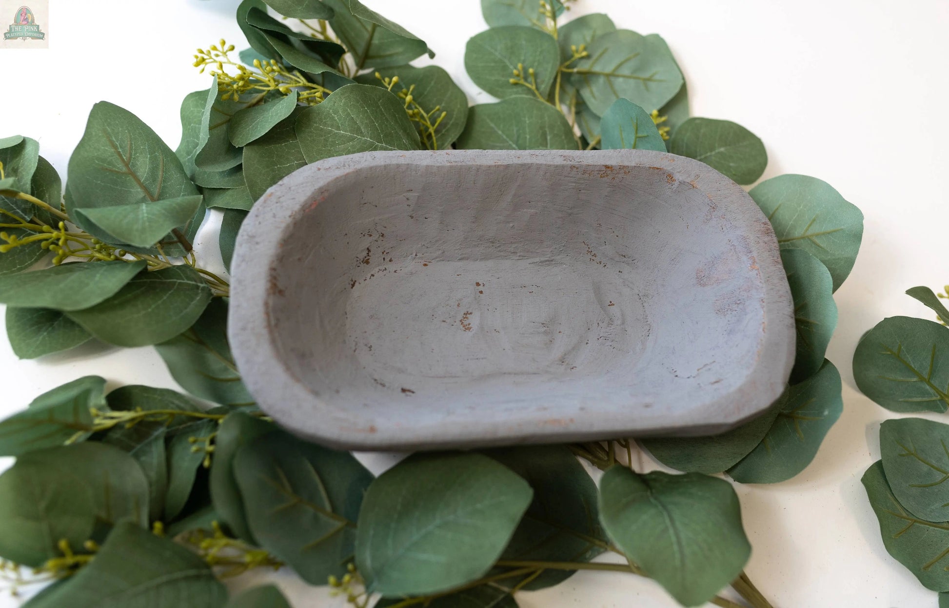 A handmade Petite Wood Bowl (9-10"), natural finish, 10x6x3 inches, from Mexico, sits empty on overlapping green eucalyptus leaves atop a white surface.