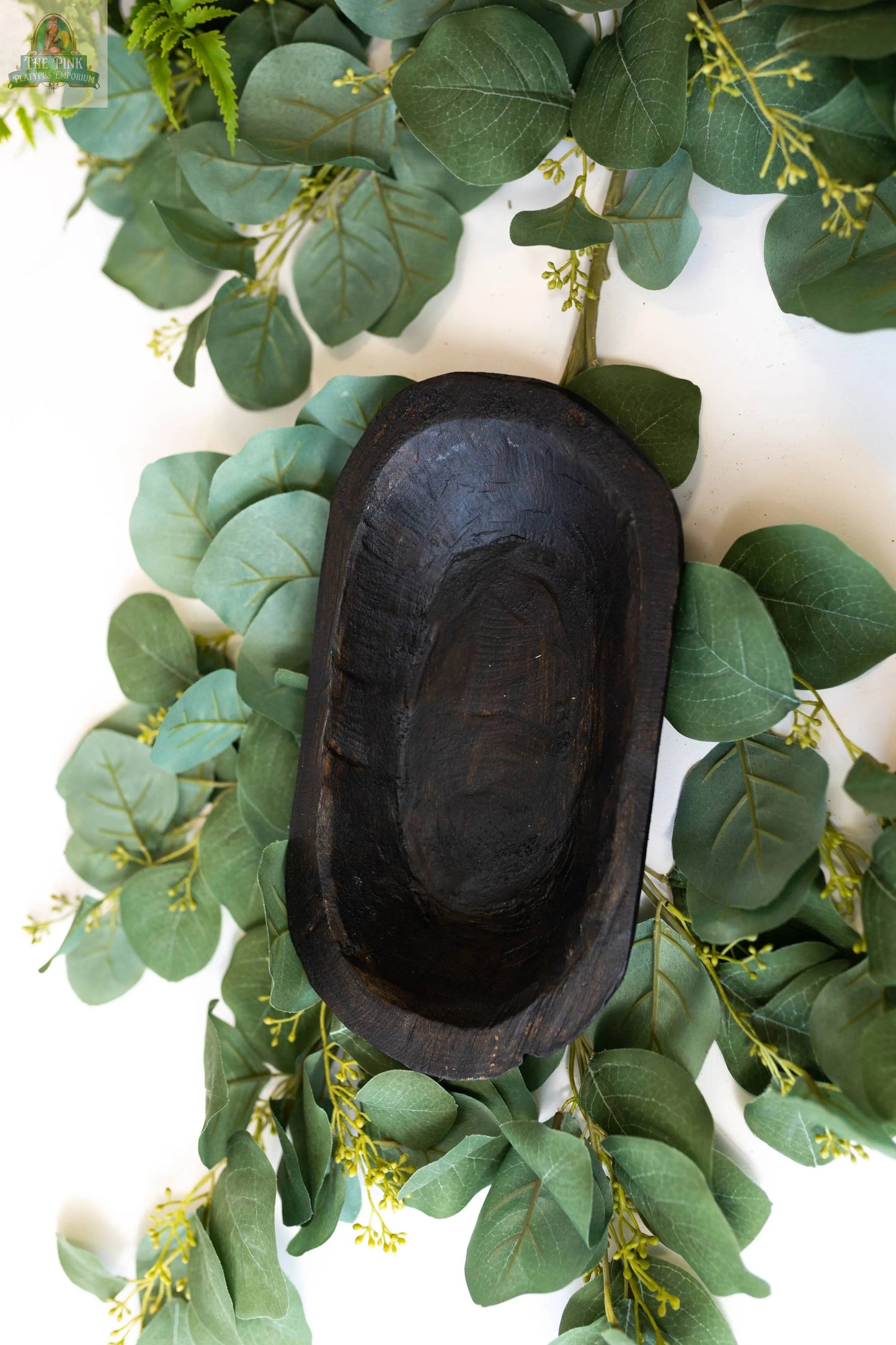The Petite Wood Bowl (9-10"), handmade in Mexico (Natural, 10x6x3), sits among eucalyptus leaves and greenery, creating a natural and rustic display.