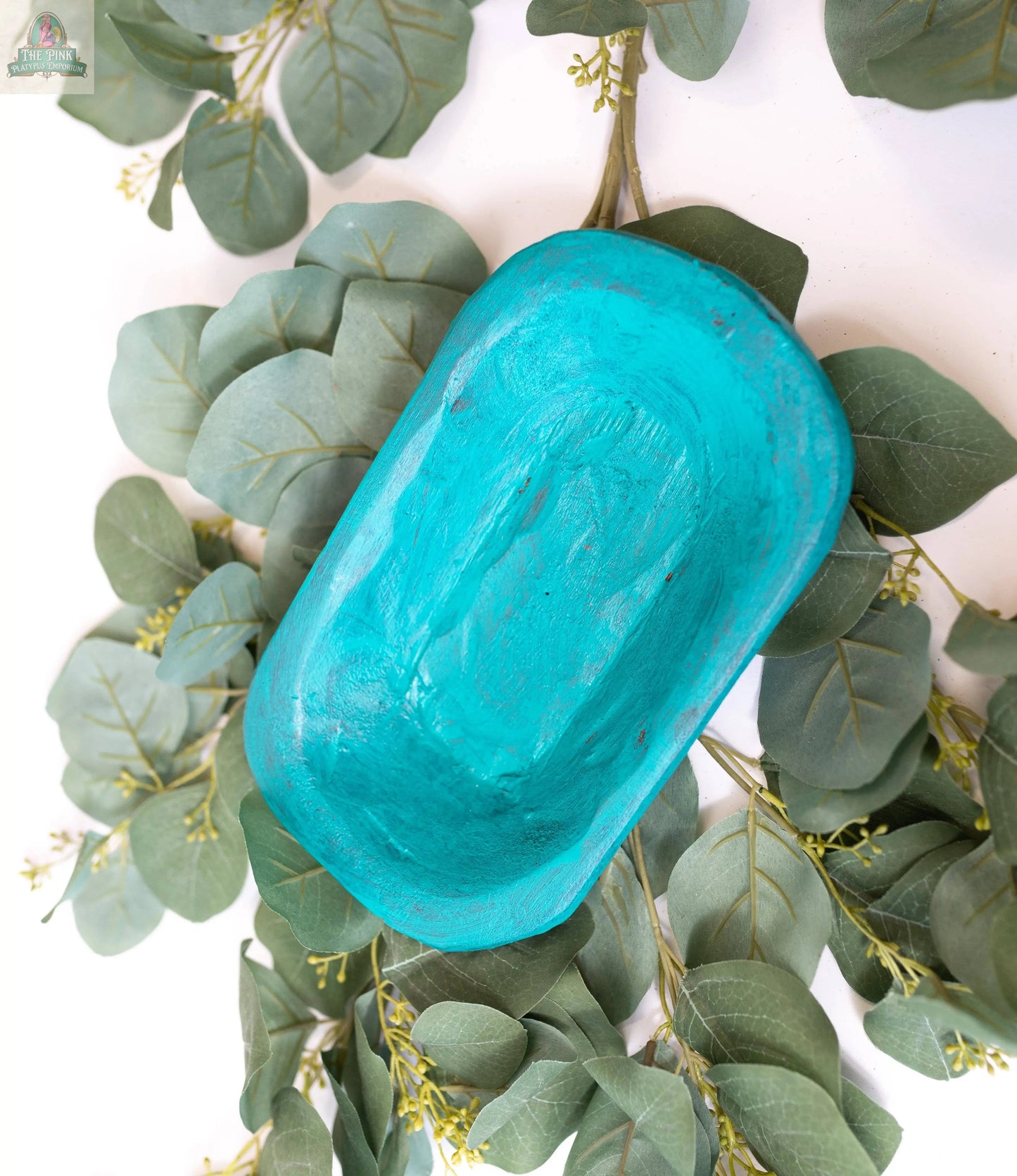 A Petite Wood Bowl (9-10"), handmade in Mexico from natural wood (10x6x3"), rests on green eucalyptus leaves against a white background, highlighting its artisanal charm and unique oval shape.