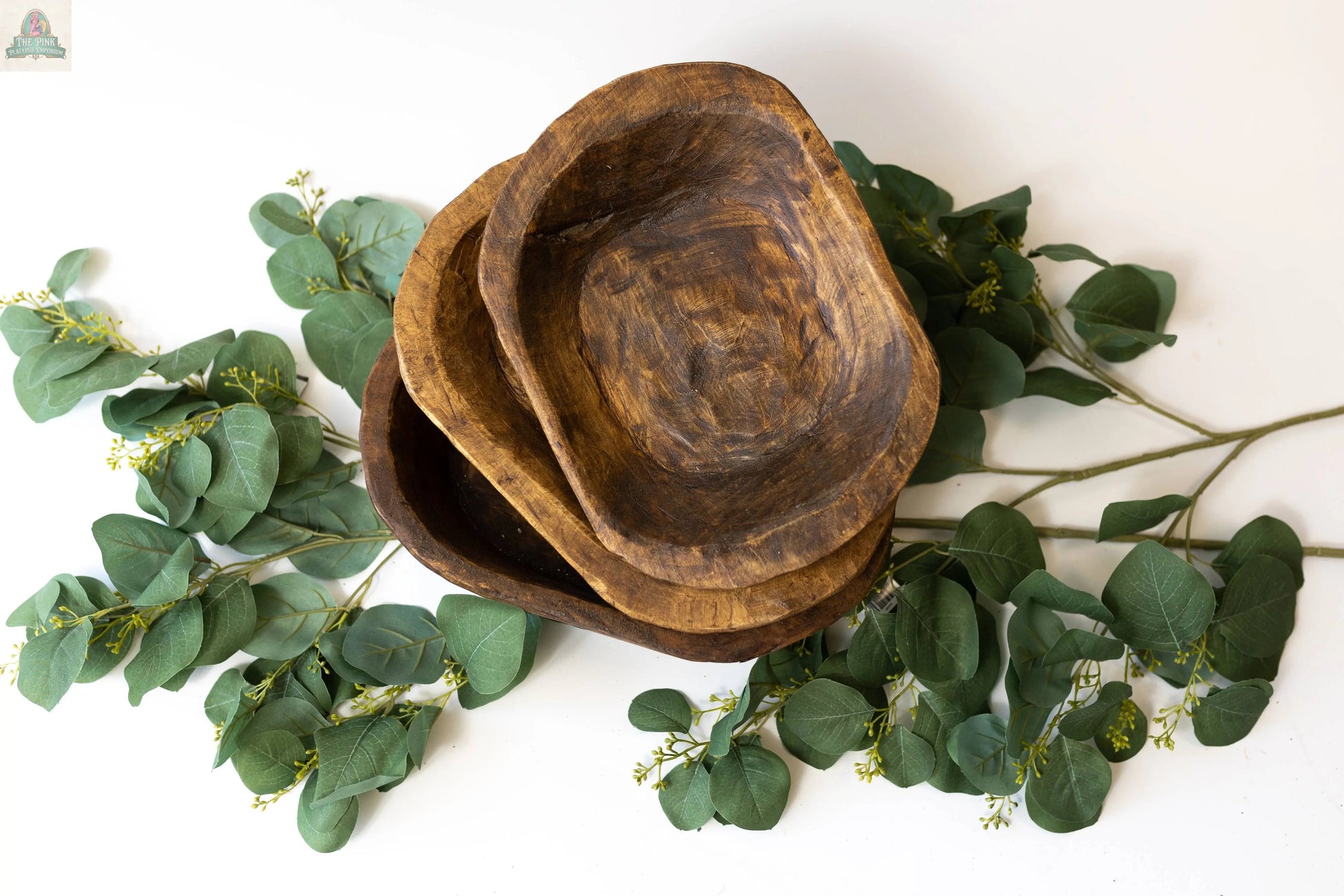 A Petite Wood Bowl (9-10"), handmade in Mexico, is stacked with others and placed on green eucalyptus branches against a white background.