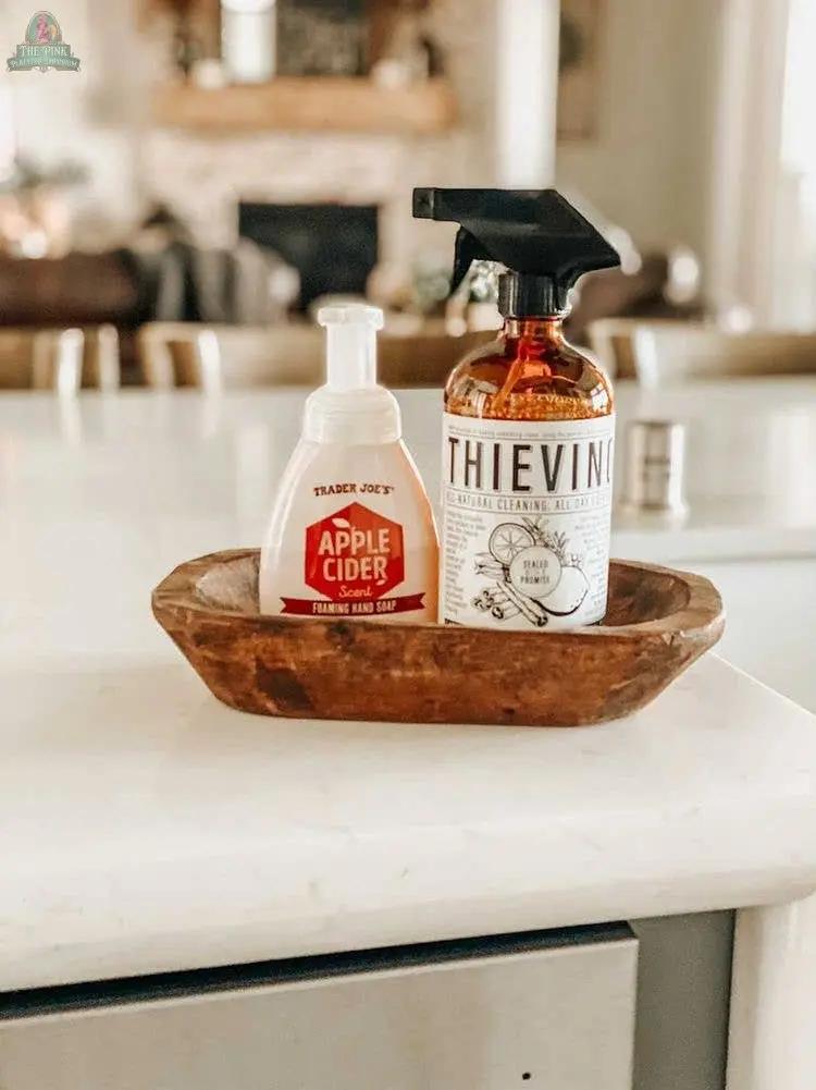 A wooden tray on a white countertop holds a Trader Joe’s Apple Cider foaming hand soap, a Thieves cleaner spray bottle, and a Candle Ready Handmade Petite Wood Bowl (#A - 7.1). A blurred kitchen is visible in the background.