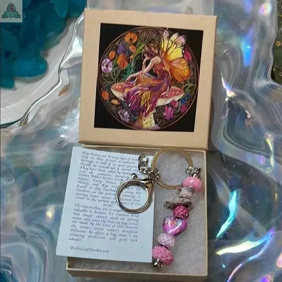 The Pink Beaded Hagstone Keychain with Fairy Gift Box includes a pink and silver hagstone keychain, a note card, and features a vibrant fairy design. It’s displayed on an iridescent wavy surface, perfect for gifting.