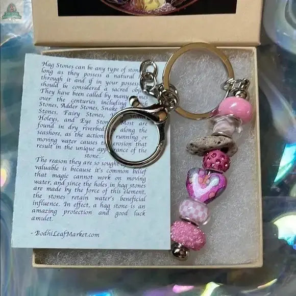 The Pink Beaded Hagstone Keychain with Fairy Gift Box features pink-patterned beads, a heart charm, and a hag stone, beautifully packaged with a note about the meaning of Sanibel Island hag stones.