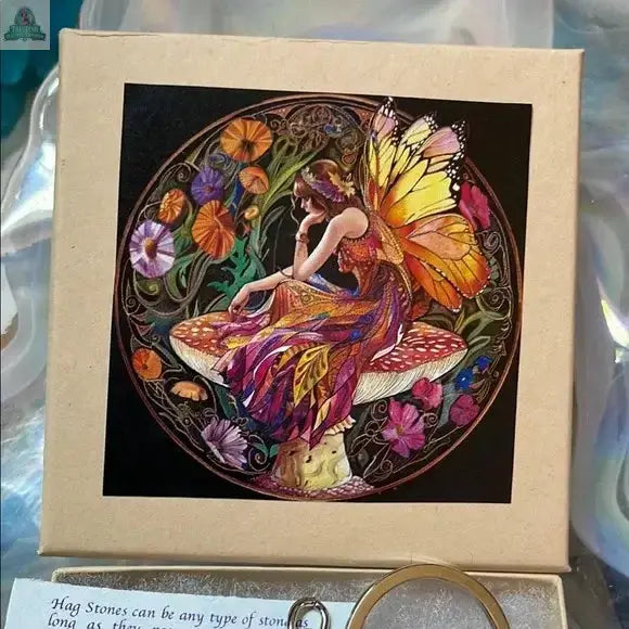 A Pink Beaded Hagstone Keychain comes in a gift box featuring a fairy with butterfly wings sitting on a mushroom, clutching a magical hagstone amid vibrant flowers and greenery, all in a circular frame.