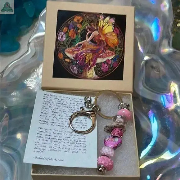 The Pink Beaded Hagstone Keychain with Fairy Gift Box features a colorful hag stone keychain, an illustrated card of a fairy on a mushroom, and a note, all set against an iridescent background that enhances its magical charm.