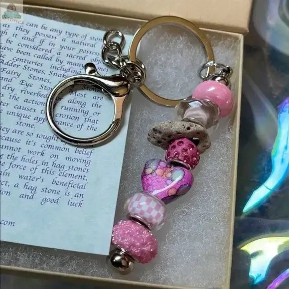 The Pink Beaded Hagstone Keychain features pink, purple, and stone beads—including a heart bead—in a fairy gift box with a card explaining the Sanibel Island-inspired magical hagstone charm and its meaning.