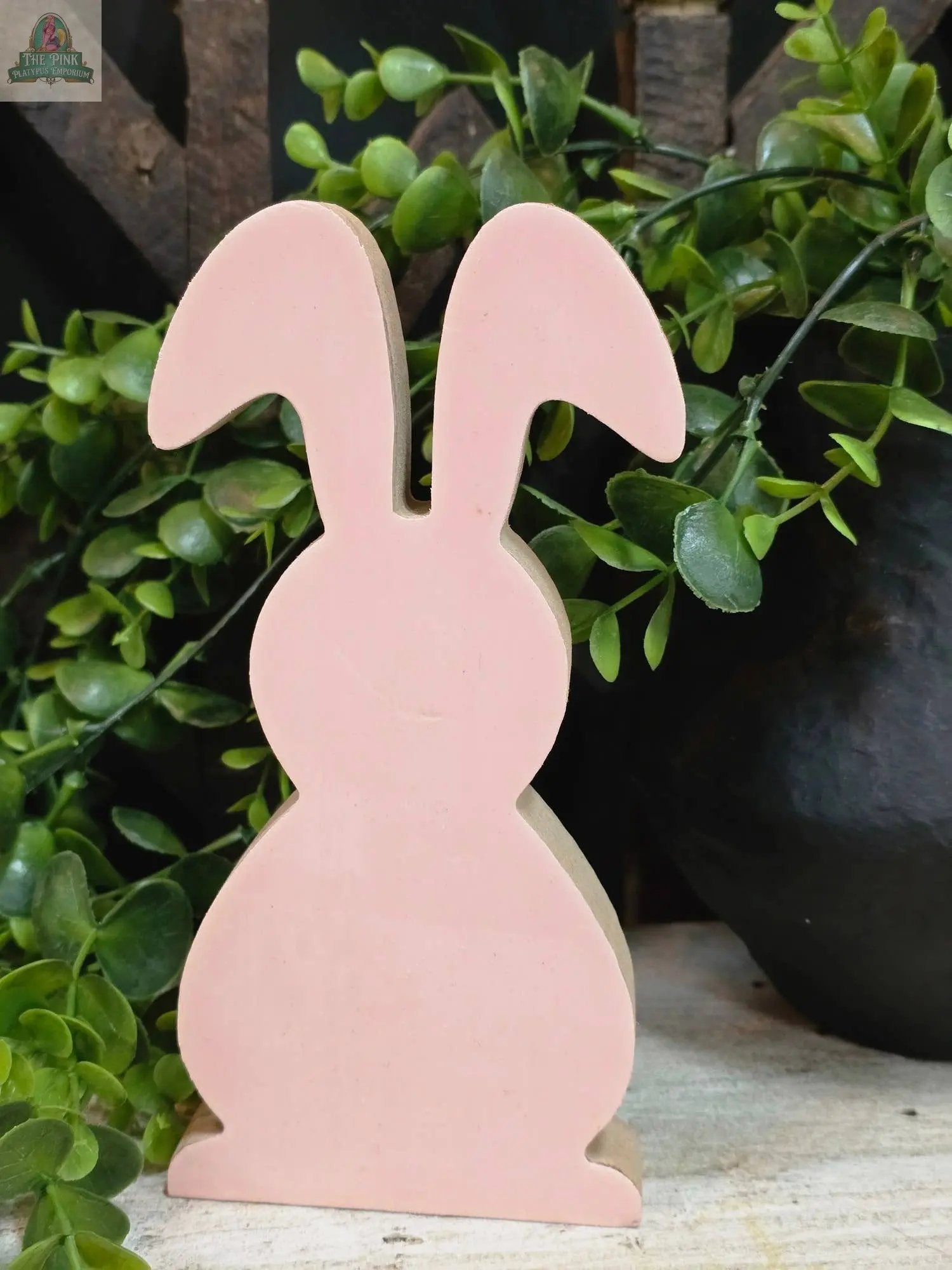 The Pink Wooden Bunny 7x3in stands upright, with green leafy plants in the background—a charming decor piece for any space.