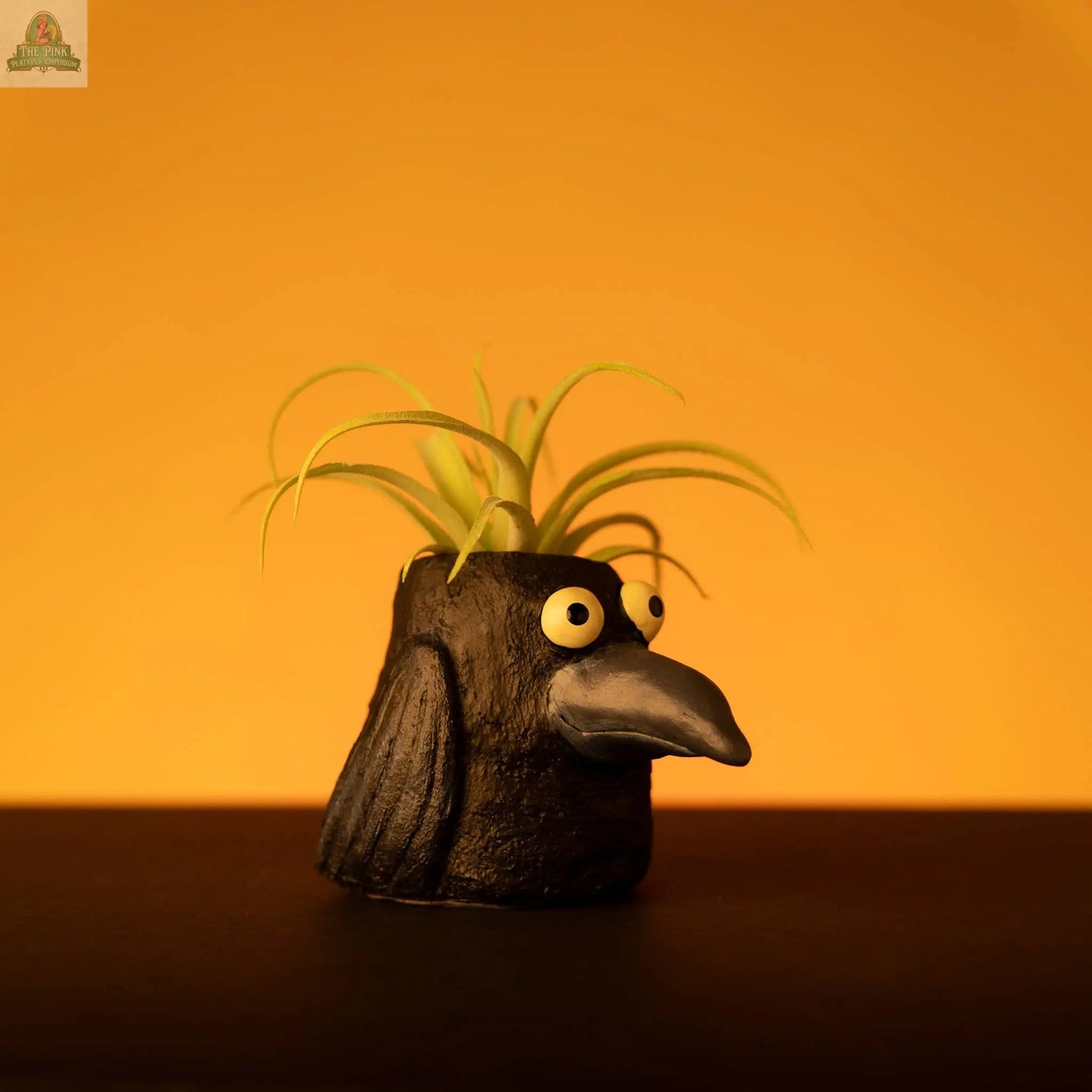 Poe the Crow Blobhouse Planter is a small black handcrafted planter shaped like a cartoonish bird with big yellow eyes and a beak, perfect for small plants and adding whimsy to your home décor. Set against a solid orange background.