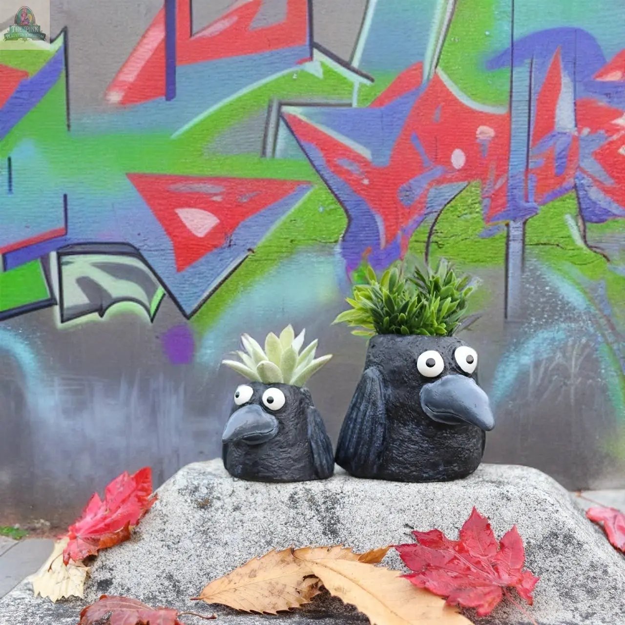 Two Poe the Crow Blobhouse Planters, shaped like black birds with googly eyes, sit on a concrete surface holding succulents. Red and brown leaves are scattered around, with colorful graffiti in the background—great for unique home décor.