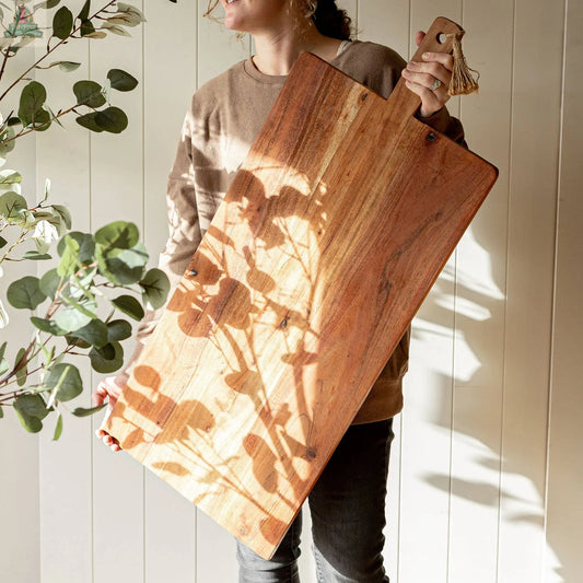 A person holds the 38 Inch Oversized Cutting Board/Charcuterie Board with plant shadows on it, standing by a light wall and leafy branches—the ideal rustic kitchen decor inspiration.