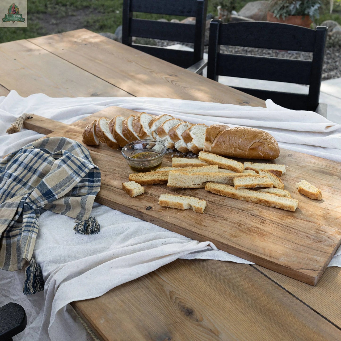 A 38 Inch Oversized Cutting Board serves sliced bread, breadsticks, and a bowl of dip outdoors. Set on an acacia charcuterie board, with a blue-beige checkered cloth nearby, black chairs, and a garden complete the scene.