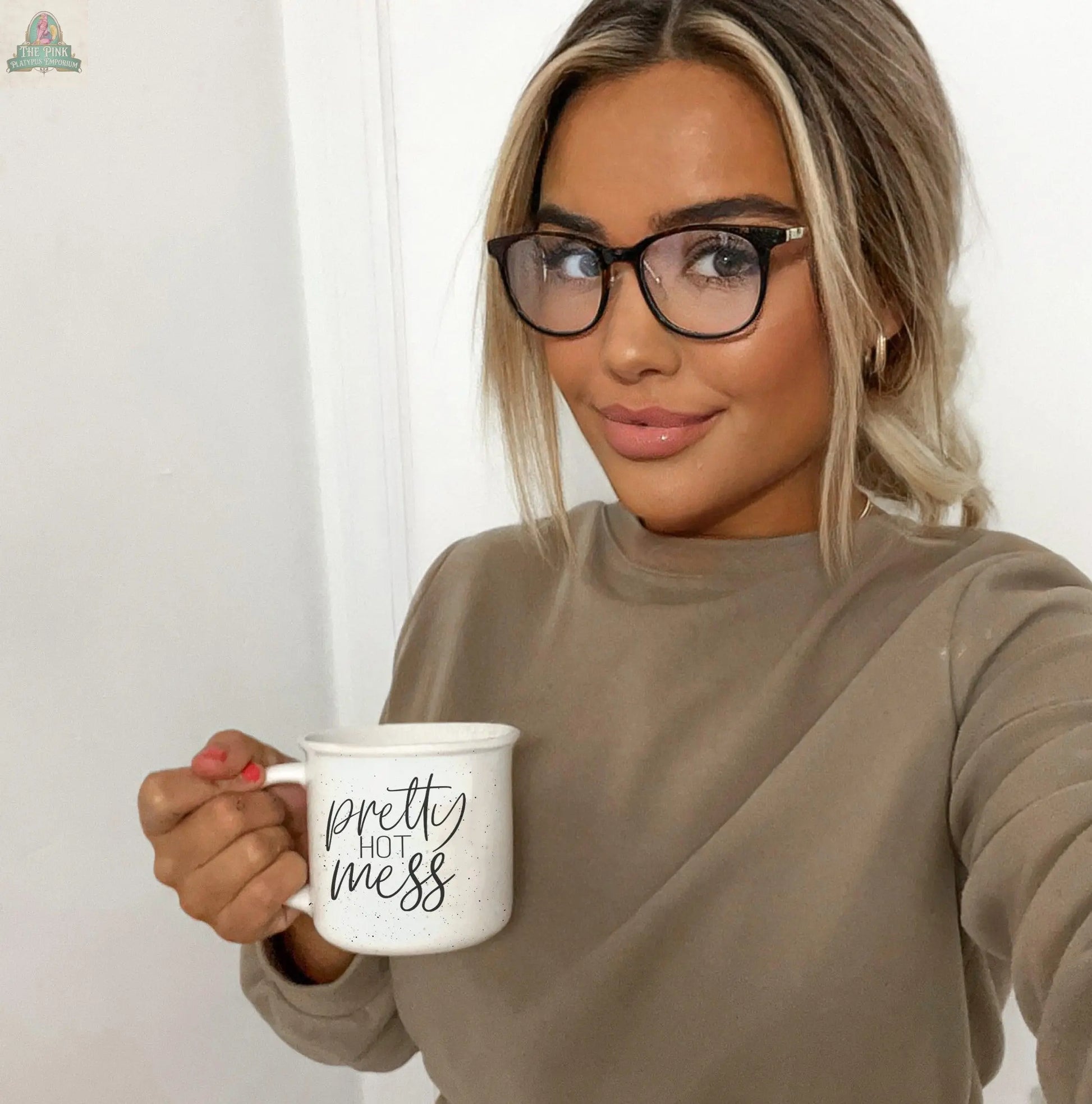 A smiling blonde woman in glasses and a taupe top holds a Pretty Hot Mess 14.5oz ceramic mug and looks at the camera.