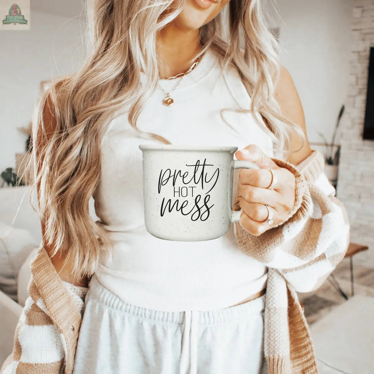 A woman with long, wavy blonde hair holds a Pretty Hot Mess 14.5oz microwave-safe mug in a bright, modern living room. She wears a white tank top, cozy cardigan, and light-colored sweatpants.
