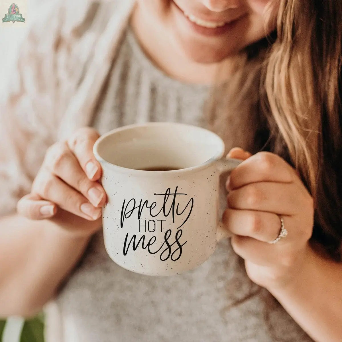 A woman with long brown hair smiles while holding the Pretty Hot Mess 14.5oz white speckled ceramic mug featuring cursive text. She wears a ring on her left hand. This microwave safe mug adds charm and practicality to your coffee routine.
