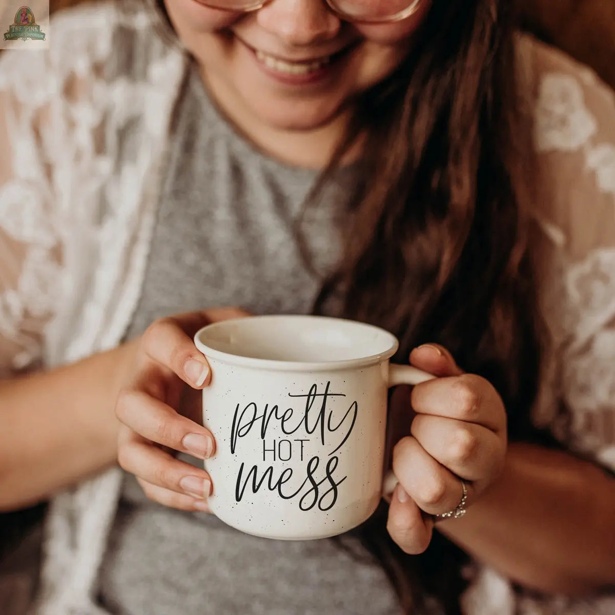 A smiling woman in glasses and a lace cardigan holds the Pretty Hot Mess 14.5oz ceramic mug, script visible. Her face is partly shown as she sits comfortably with both hands around the microwave safe mug.
