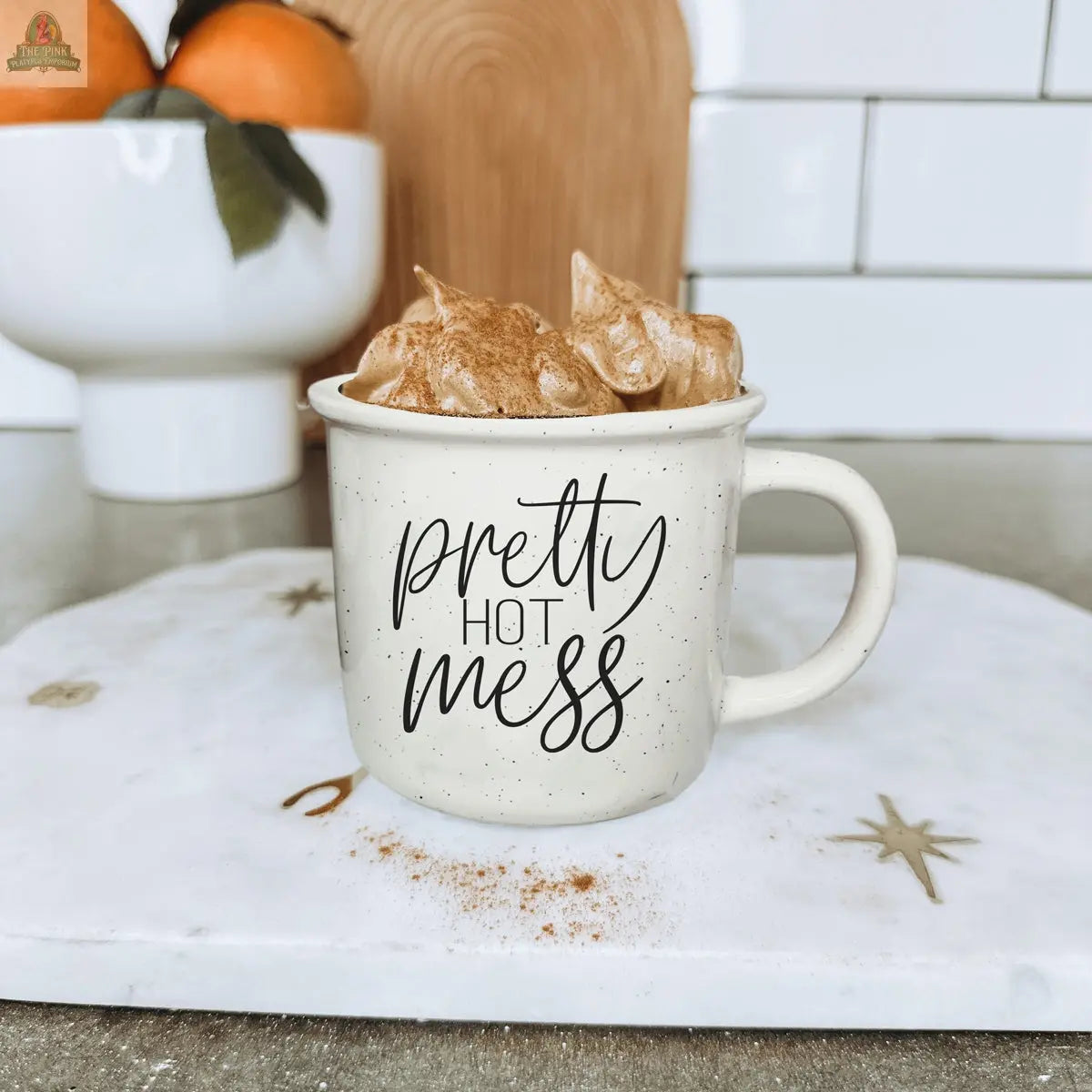 A Pretty Hot Mess 14.5oz beige ceramic mug sits on a white marble surface, filled with a creamy, swirled drink topped with cinnamon. In the background, oranges in a white bowl and a wood cutting board are visible.