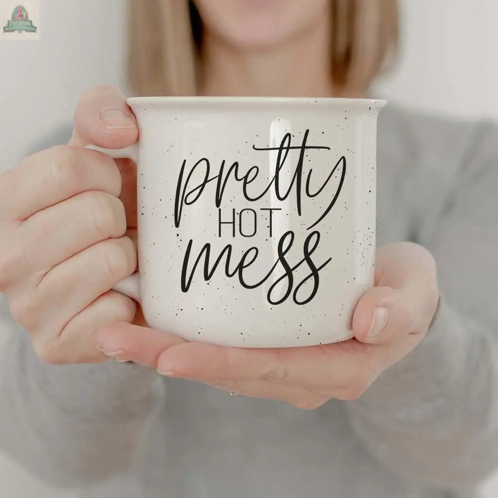 A woman in a grey shirt holds the Pretty Hot Mess 14.5oz mug, a white speckled ceramic cup labeled "pretty hot mess" in bold script. It's a stylish, microwave-safe mug perfect for daily use.