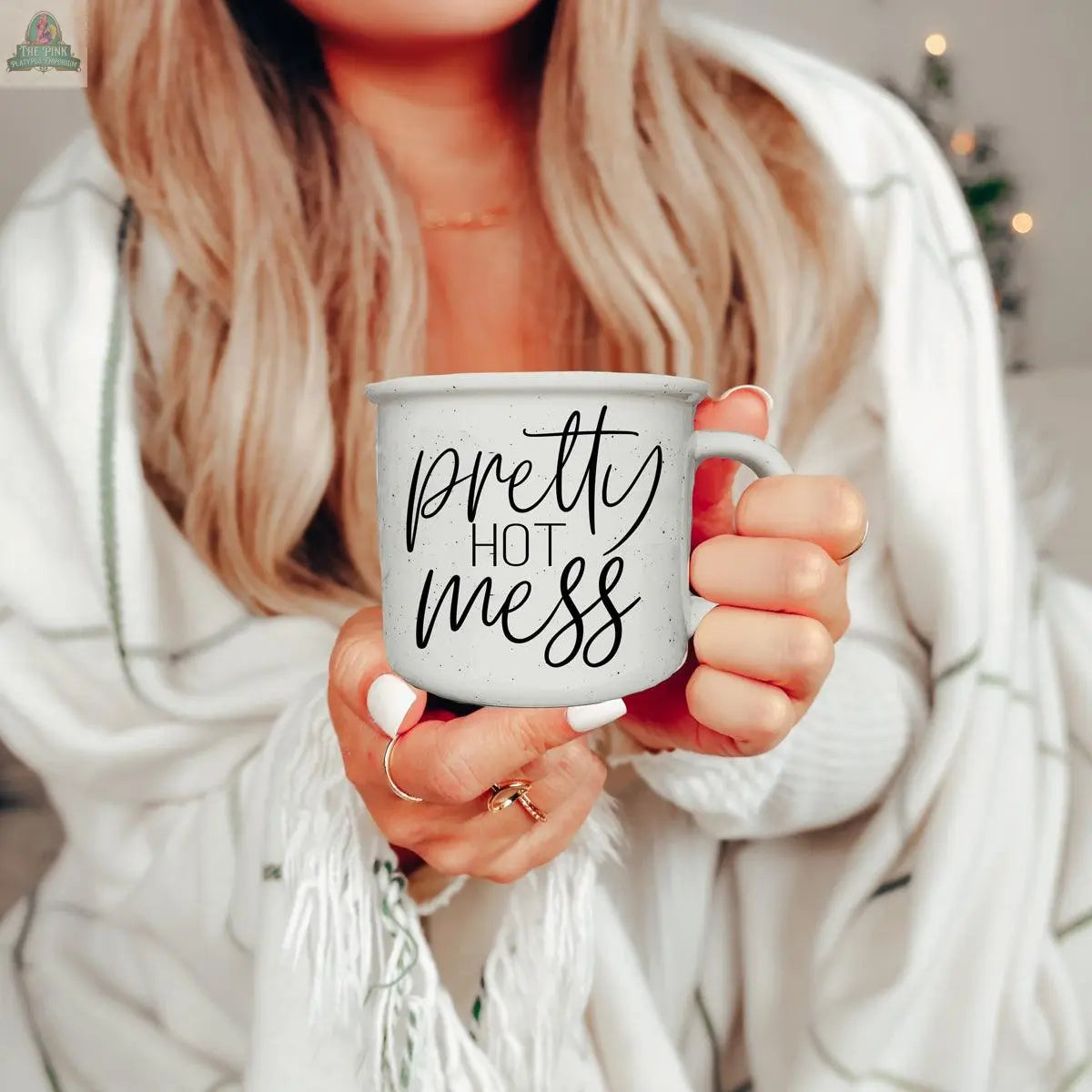A woman with long blonde hair and white nails holds the Pretty Hot Mess 14.5oz ceramic mug with black script, wrapped in a white and green striped blanket. The mug is both microwave and dishwasher safe.