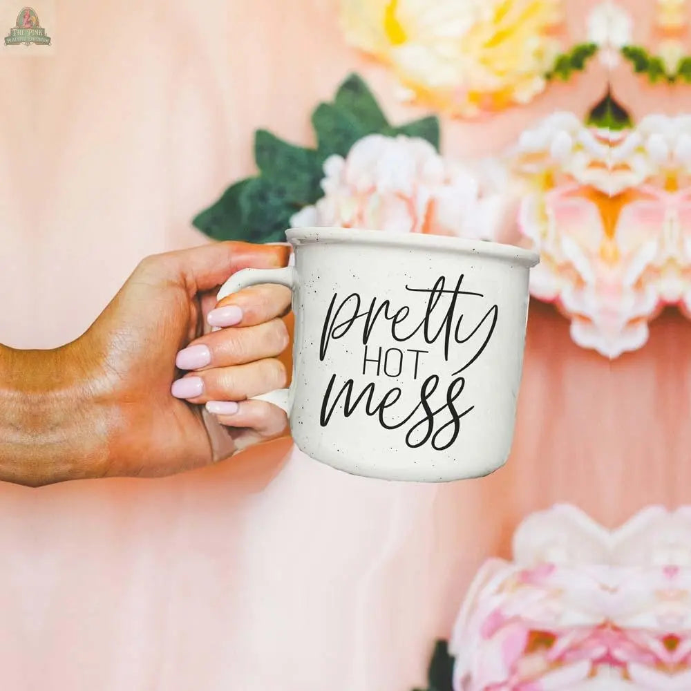 A hand with light pink nails holds the Pretty Hot Mess 14.5oz white ceramic mug with “pretty hot mess” in script font, against a blurred background of soft pink and white flowers.