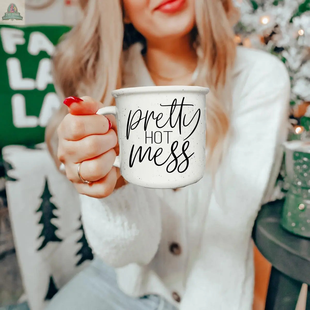 A woman in a white sweater holds the Pretty Hot Mess 14.5oz ceramic mug, which features “pretty hot mess” text. Surrounded by holiday décor and green pillows with tree designs, the mug is microwave and dishwasher safe.