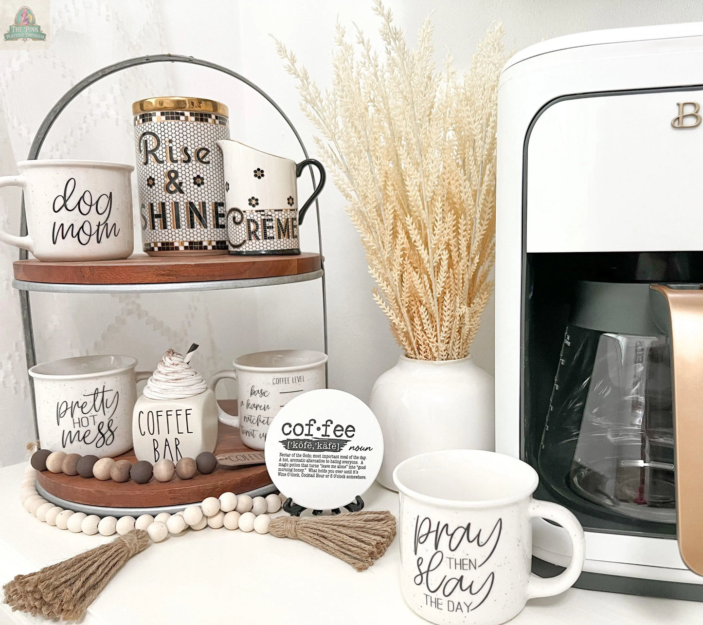 A coffee station features a white coffee maker, a tiered stand with the Pretty Hot Mess 14.5oz ceramic mug, mugs labeled “dog mom,” a “coffee bar” canister, and is decorated with beads and dried grass.