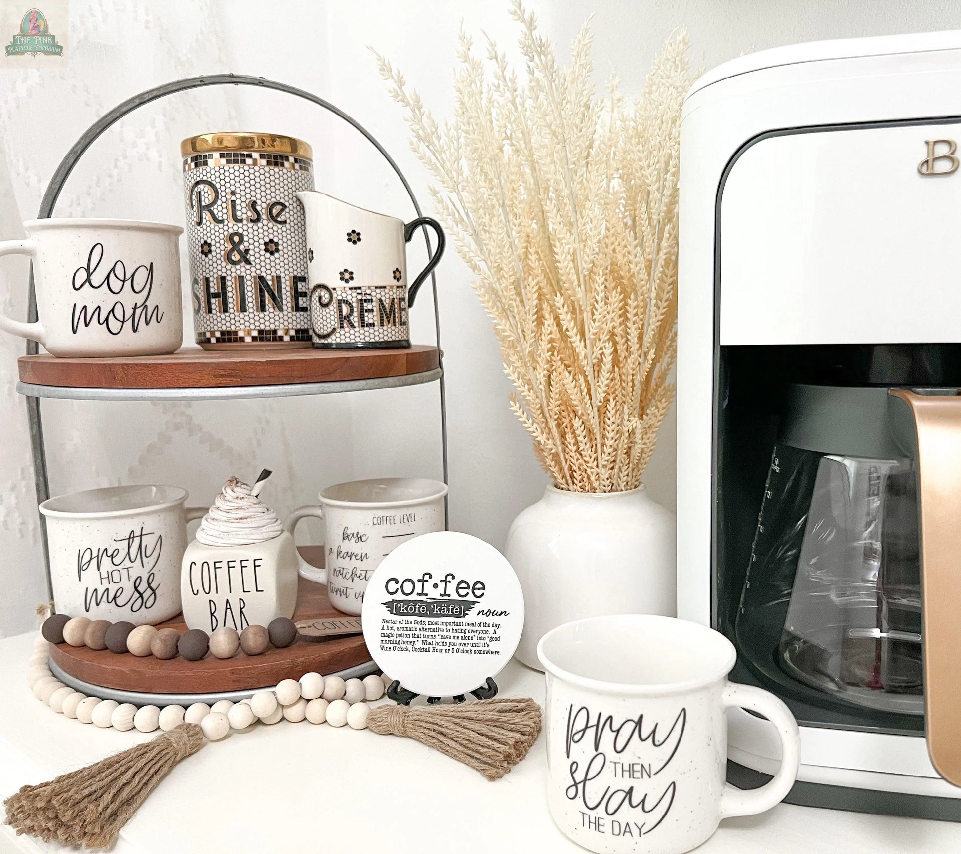A coffee station features a white coffee maker, a tiered stand with the Pretty Hot Mess 14.5oz ceramic mug, mugs labeled “dog mom,” a “coffee bar” canister, and is decorated with beads and dried grass.