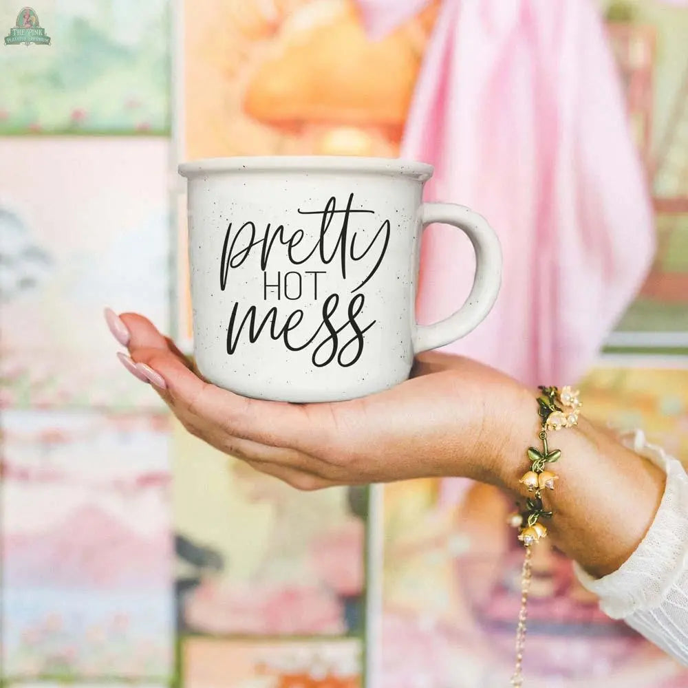 A hand holds the Pretty Hot Mess 14.5oz white ceramic mug with “pretty hot mess” in cursive. In the background are vibrant, blurred paintings and a person in pink with a floral bracelet. This mug is microwave safe.