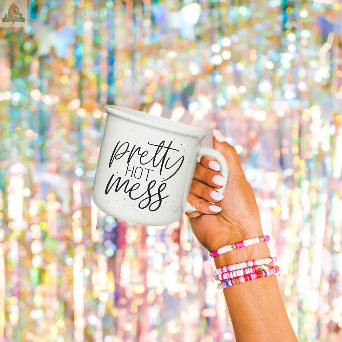 A hand with colorful beaded bracelets holds the Pretty Hot Mess 14.5oz white ceramic mug against a shiny, sparkly background. This microwave-safe mug is perfect for your daily coffee rituals.