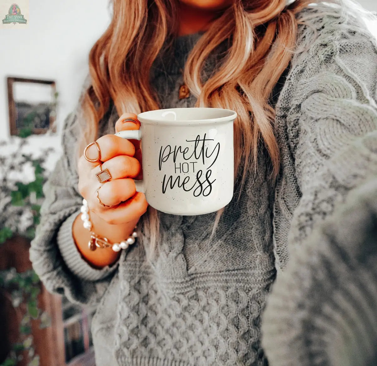 A woman in a chunky gray sweater holds the Pretty Hot Mess 14.5oz ceramic mug, adorned with cursive text. She wears multiple rings and bracelets, with greenery blurred behind her. The mug is both dishwasher and microwave safe.