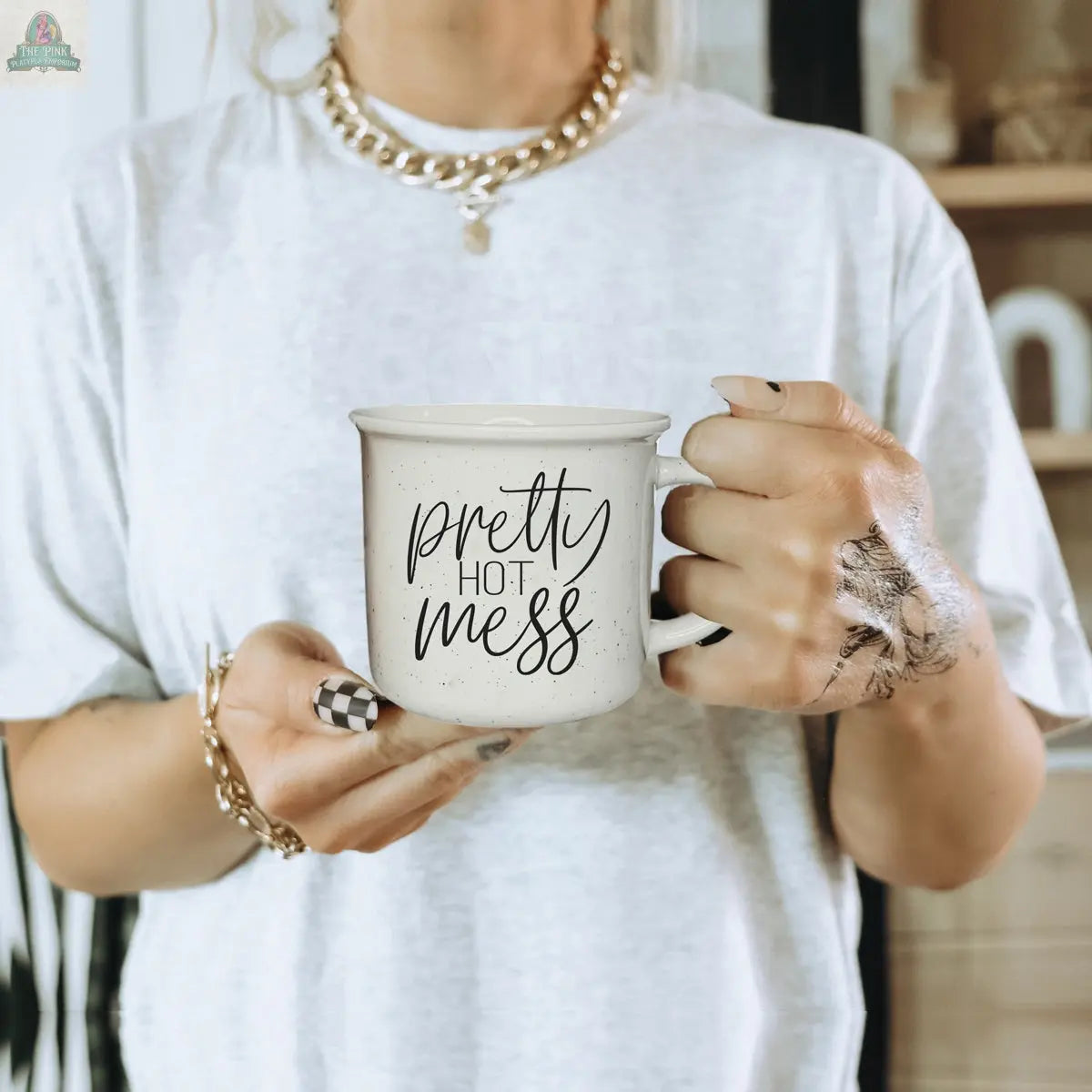 A person in a gold chain and white tee holds the Pretty Hot Mess 14.5oz ceramic mug with checkered nail art and ink-stained hands. The softly blurred background highlights the mug, which is both dishwasher and microwave safe.