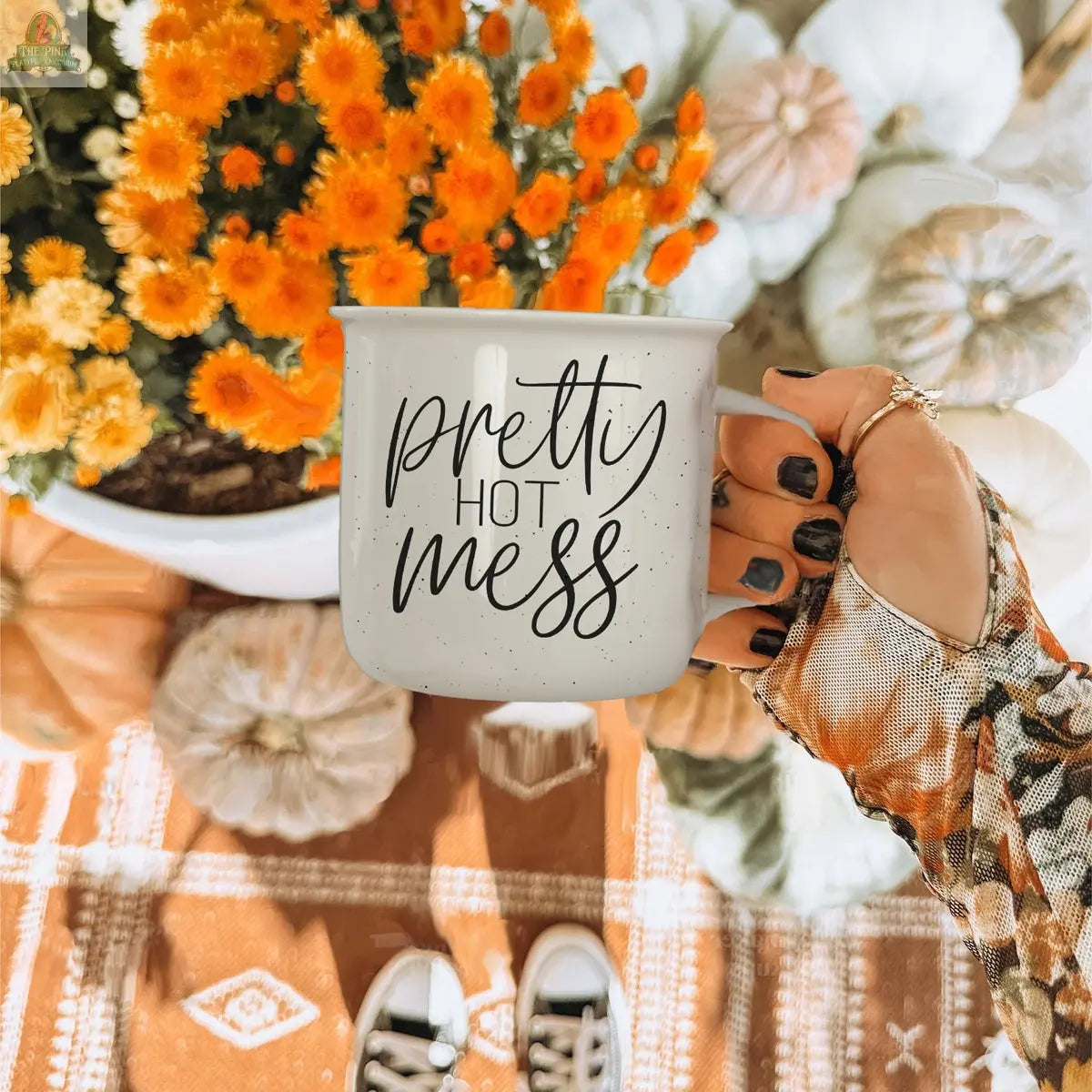 A hand with black nail polish holds a microwave-safe Pretty Hot Mess 14.5oz ceramic mug over an autumn scene with orange flowers, white pumpkins, a patterned rug, and partially visible sneakers.