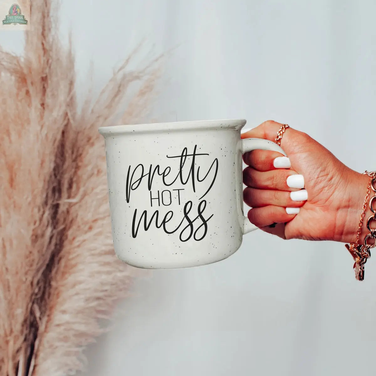 A hand with white nails and gold rings holds the Pretty Hot Mess 14.5oz white speckled ceramic mug, featuring cursive text, with beige pampas grass in the background; this microwave-safe mug is perfect for your favorite drinks.