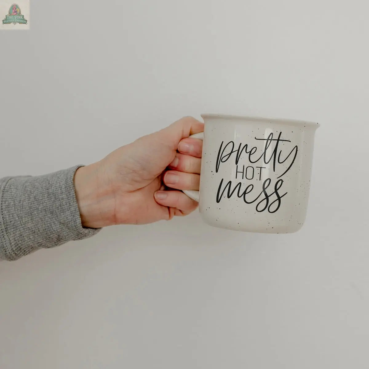 A hand in a grey sleeve holds the Pretty Hot Mess 14.5oz white ceramic mug with black cursive text, set against a plain light background.