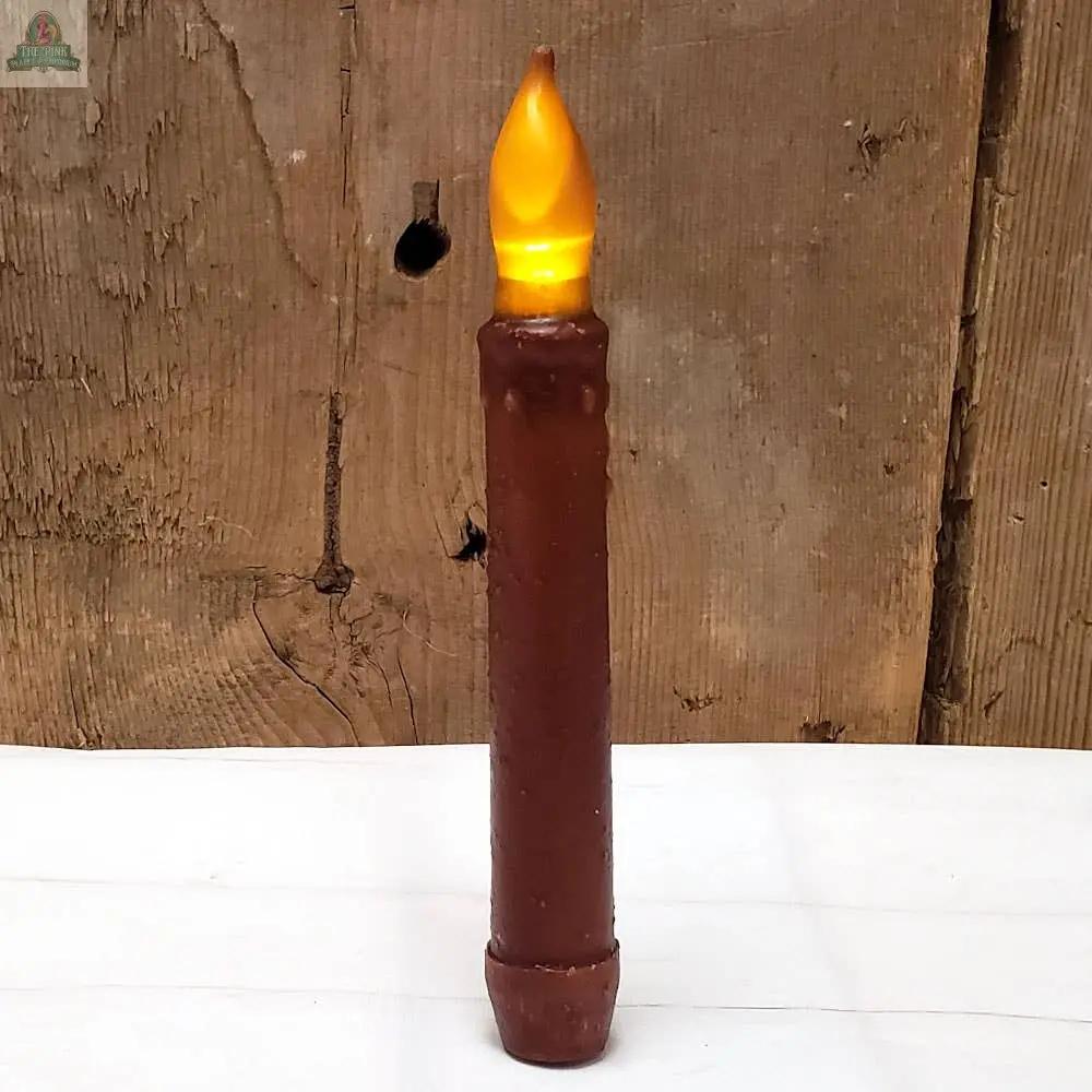 The Primitive Mustard LED Taper w/ Timer, featuring a battery-operated yellow flame-shaped light, stands upright on a white surface against a rustic wooden background.