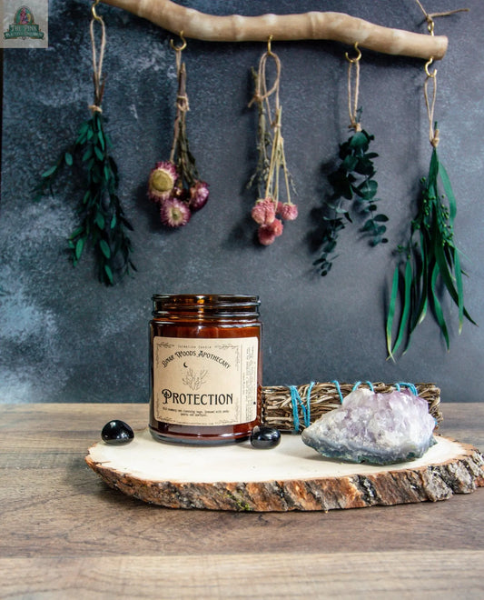 A brown glass jar labeled "Protection" sits on a wooden slab with black tourmaline, a crystal, and sage. Dried flowers and herbs hang in the background, adding to the metaphysical mood.