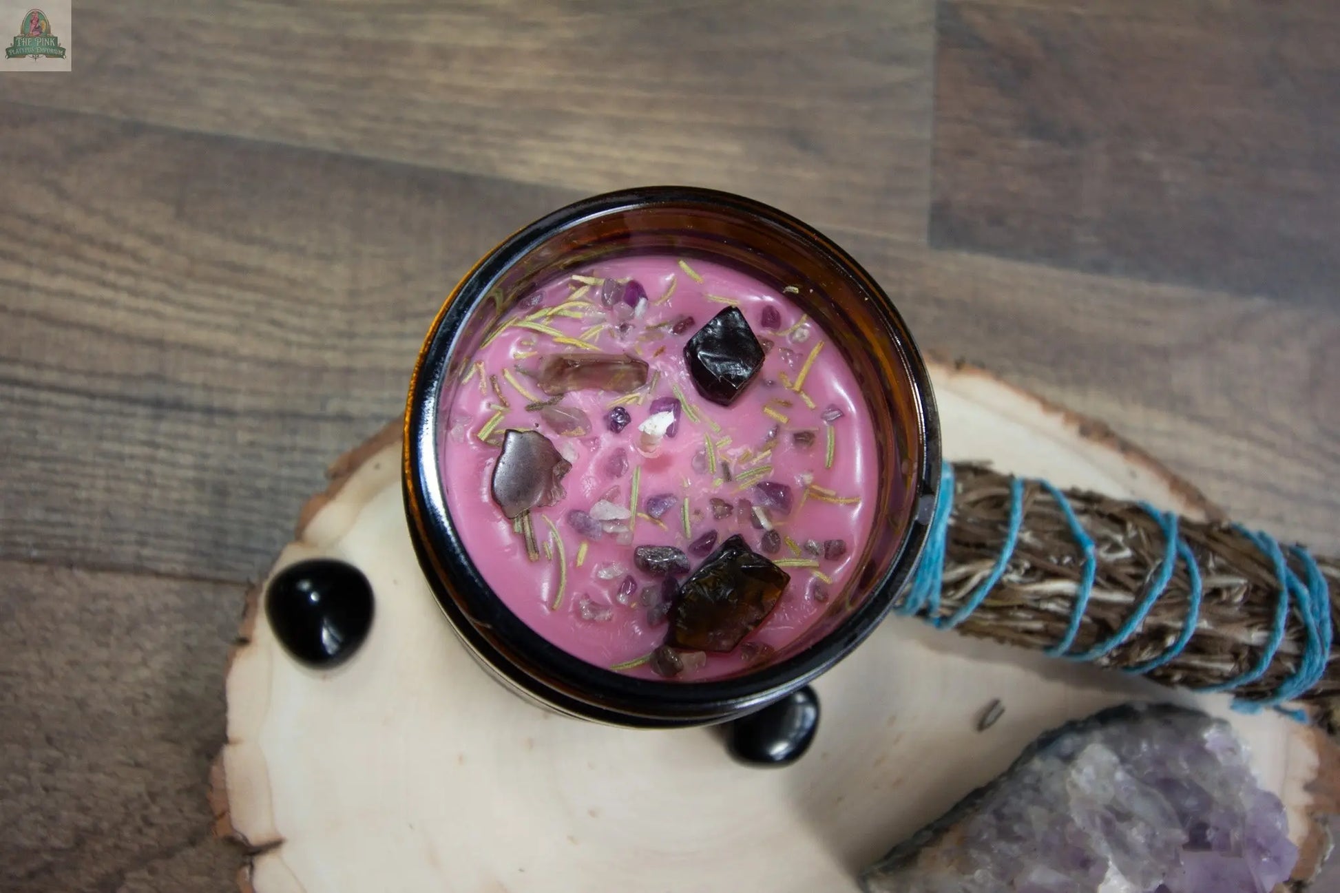 Protection, a pink candle in a glass jar adorned with crystals and dried herbs, sits on a wooden tray alongside sage, black tourmaline, and amethyst—ideal as a metaphysical mood booster for any space.