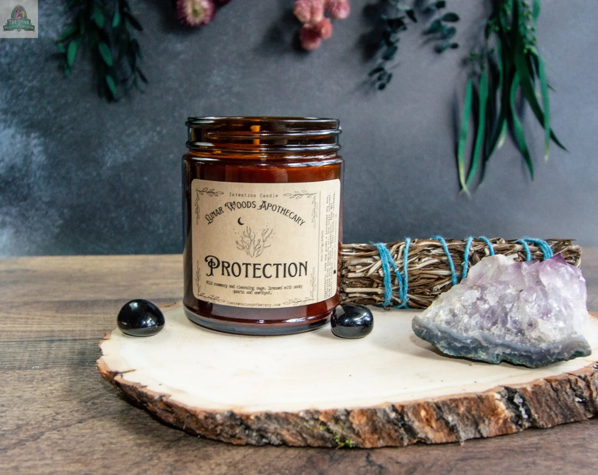 The "Protection" candle, in brown glass, sits on a wooden slab with black tourmaline stones, amethyst, and sage. Greenery and flowers blur in the background, creating a metaphysical scene for mood boosting and spiritual cleansing.