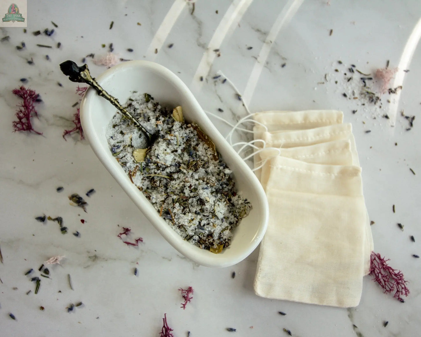 A white dish holds Protection, a blend of salt and dried herbs, on marble with a small spoon inside. Four empty drawstring muslin bags and scattered herbs hint at your next soothing herbal bath ritual.