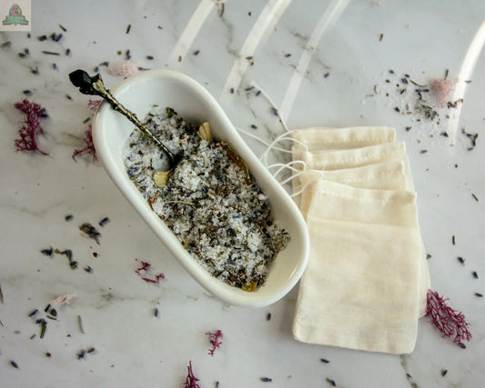 A white dish holds Protection, a blend of salt and dried herbs, on marble with a small spoon inside. Four empty drawstring muslin bags and scattered herbs hint at your next soothing herbal bath ritual.