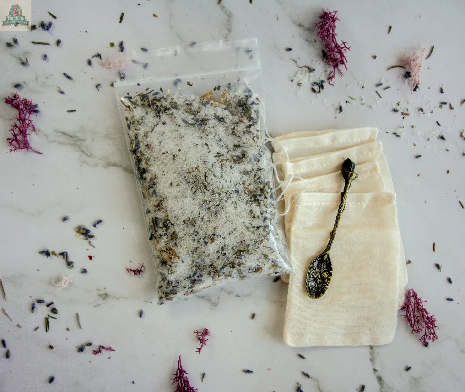 A clear bag of "Protection" bath salts with dried herbs, a small ornate metal spoon, and two empty white muslin drawstring bags are arranged on marble—ideal for making your own protective bath tea or soak among scattered dried flowers and herbs.